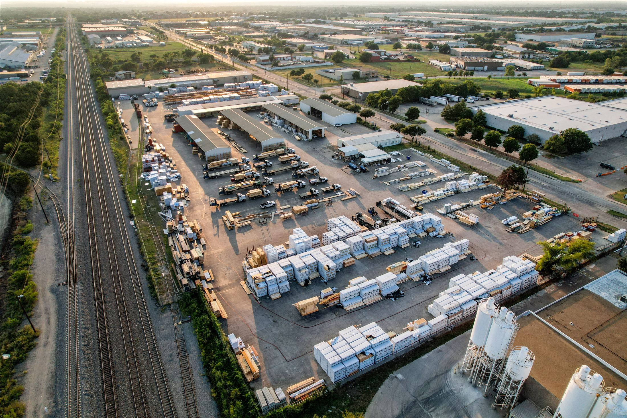 Aerial view of a building-materials yard with flatbed trucks, lumber storage, and open-air sheds